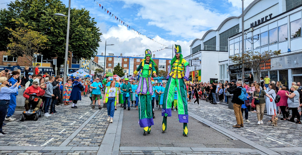 Stilt walkers and the carnival parade with people looking on from the sides of the road